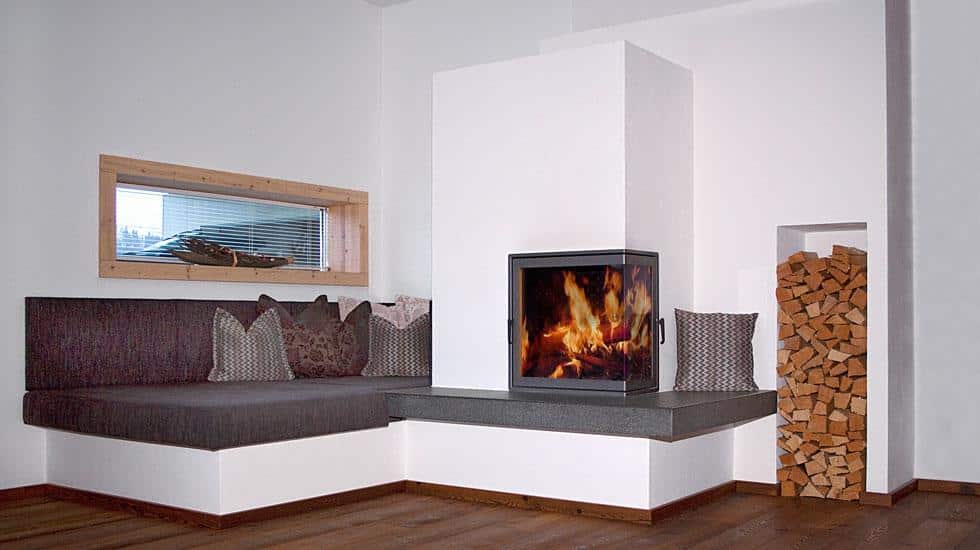 Eckkamin mit großer Sichtscheibe, integrierter Holzbank und Sitzecke in modernem Wohnzimmer mit Holzlager