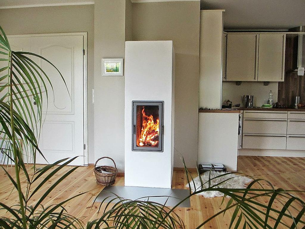 Schlichter Kaminofen mit Hochformat-Feuersicht in offenem Wohnraum mit Holzboden und Küche im Hintergrund