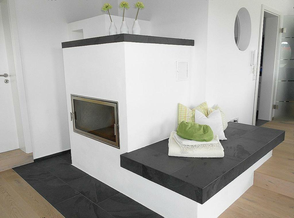 Weißer Kaminofen mit großer Sitzbank und dunkler Natursteinabdeckung in modernem, hellen Wohnraum